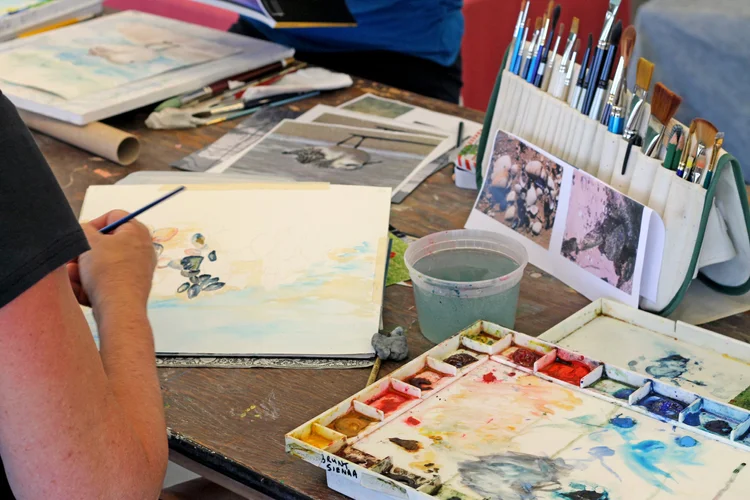 An adult participant paints a watercolor scene at a workshop table filled with brushes, paint palettes, reference photos, and art supplies.