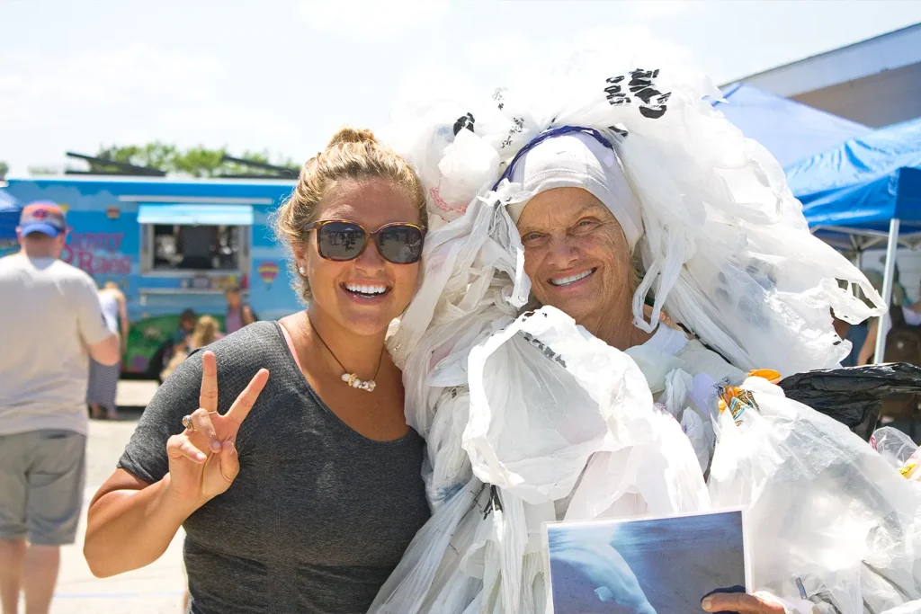 Two smiling women pose together at Barnegat Bay Day, one dressed in an outfit made entirely of plastic bags to raise awareness about pollution.