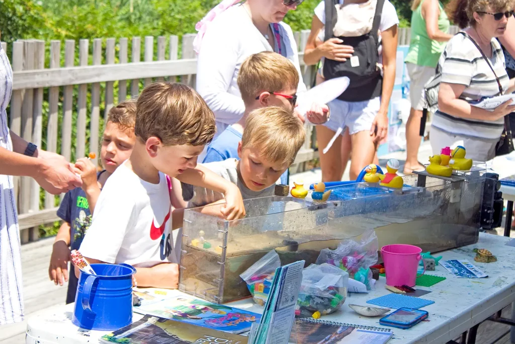 Children gather around a water table experiment at Barnegat Bay Day, learning about tides and watersheds through hands-on play with rubber ducks and sand.
