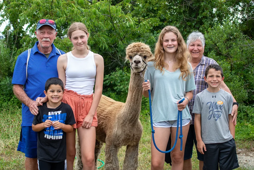 A smiling family poses with an alpaca at Barnegat Bay Day, standing on a grassy field surrounded by lush green trees.