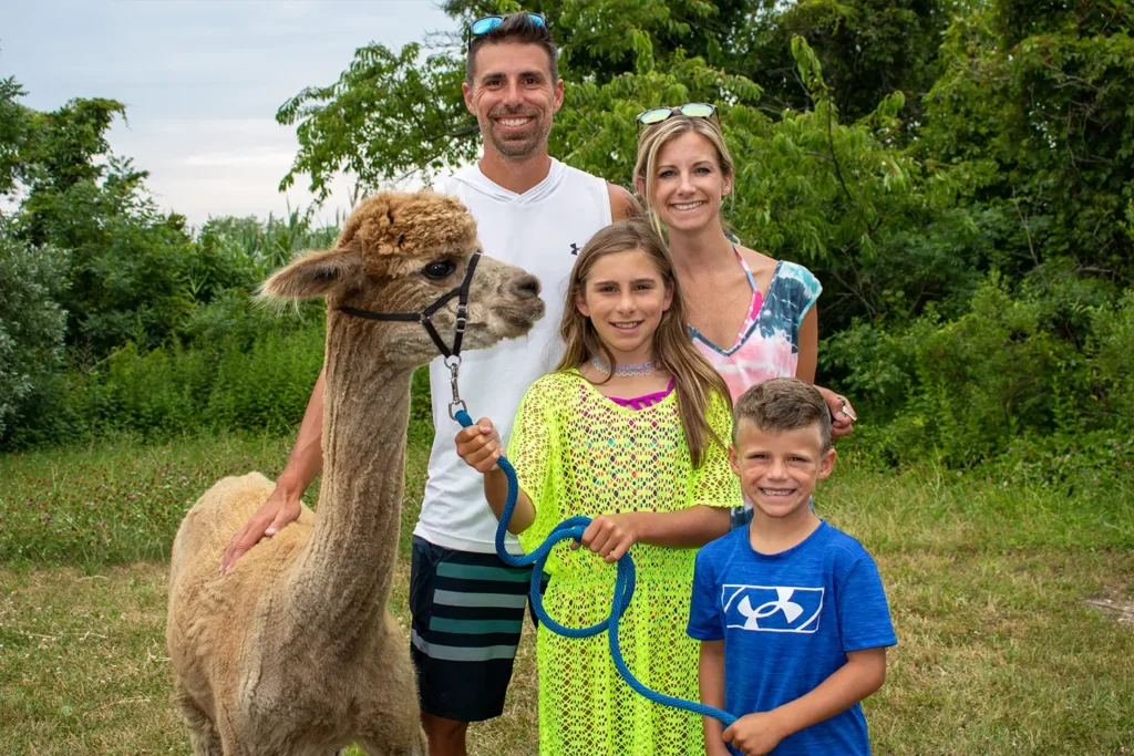 A smiling family poses with an alpaca at Barnegat Bay Day, standing on the grass with trees in the background.