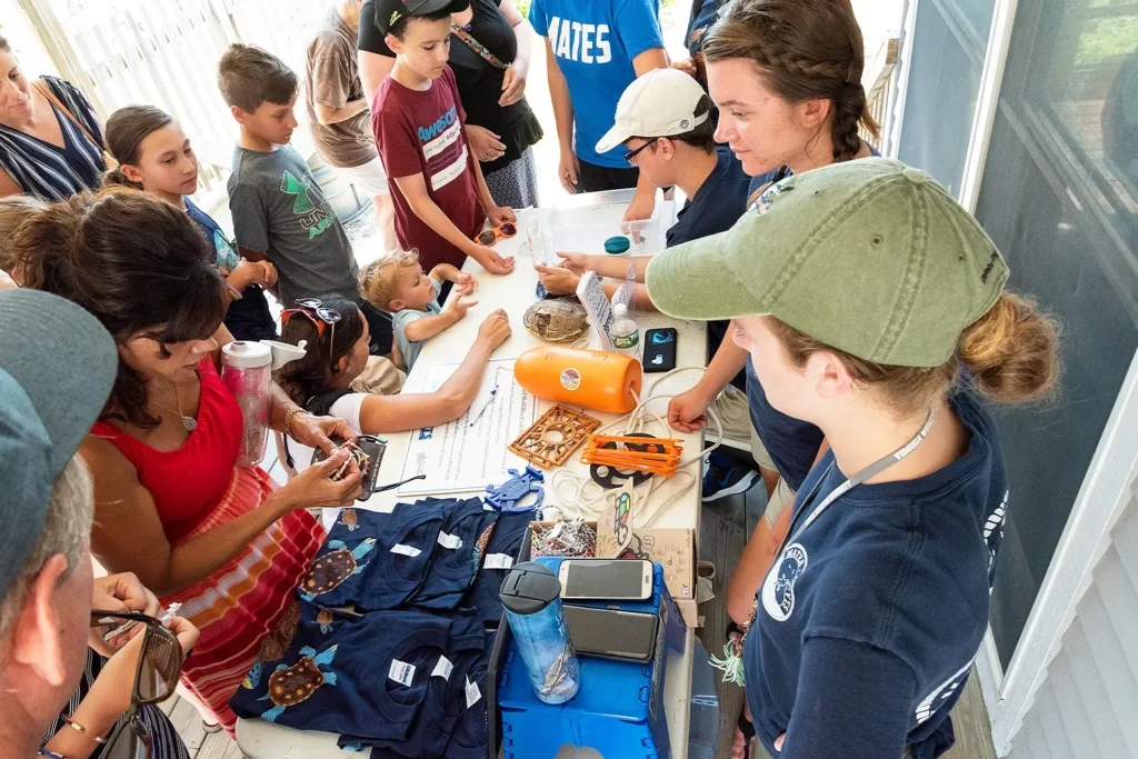 A group of children and adults gather around an educational table display, examining marine science tools and turtle shells at Barnegat Bay Day.