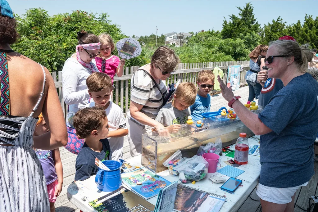 Children and families gather around an outdoor water flow table as a staff member demonstrates with rubber ducks at Barnegat Bay Day.