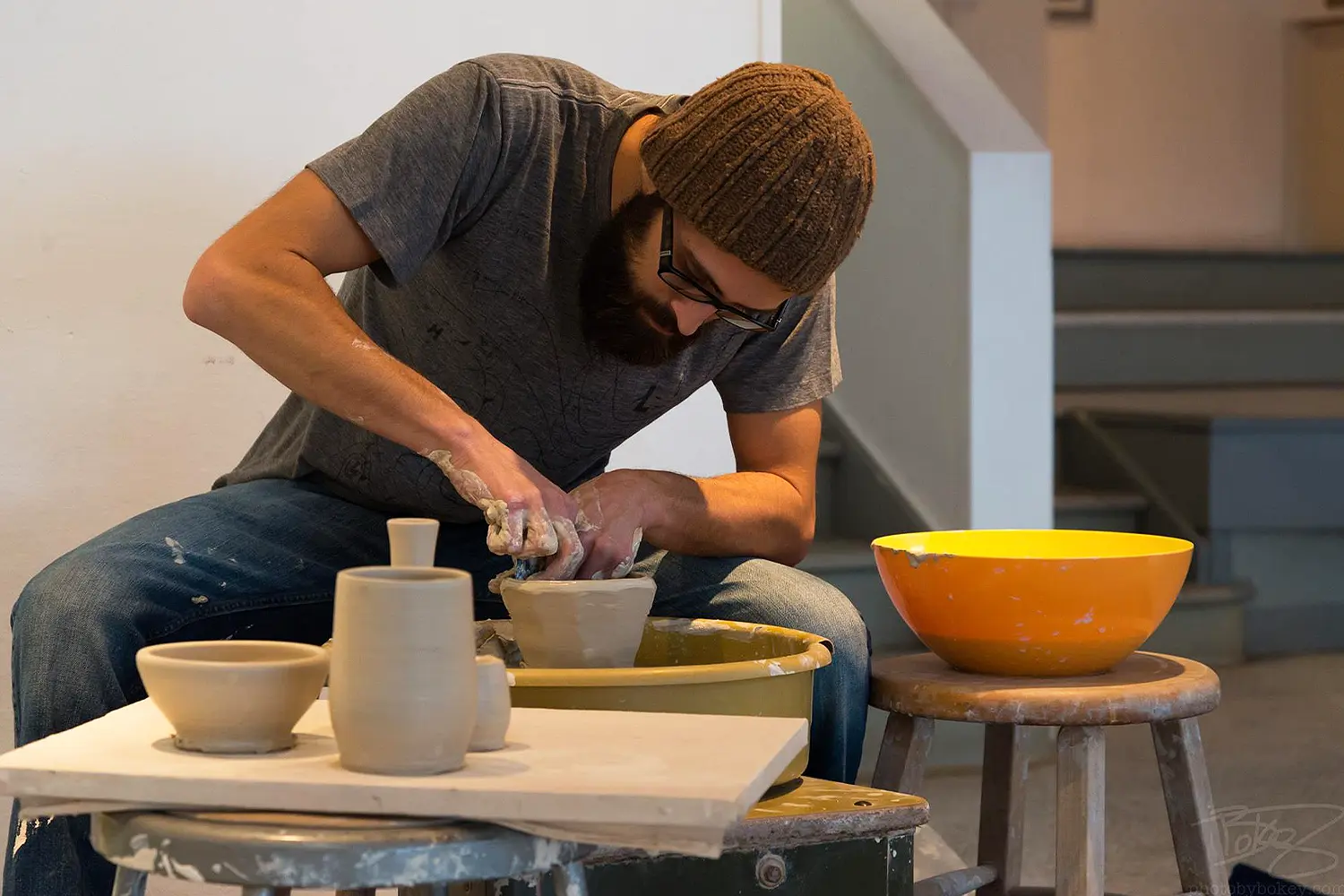 An artist in a knit hat and glasses shapes a clay bowl on a pottery wheel during a ceramics session.