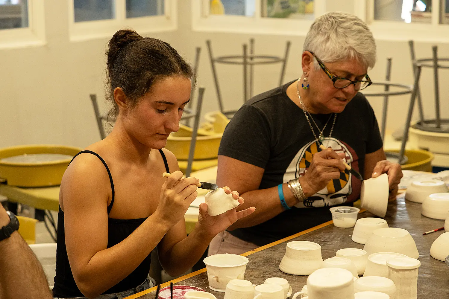 Two people paint glaze onto handmade ceramic cups in a pottery studio.