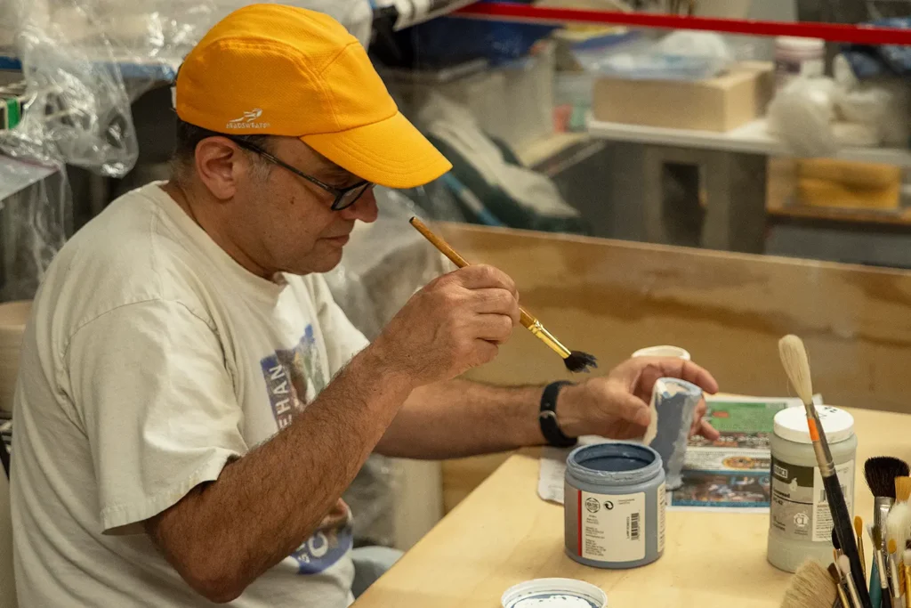 Person wearing an orange hat paints glaze onto a ceramic cup at a studio table.
