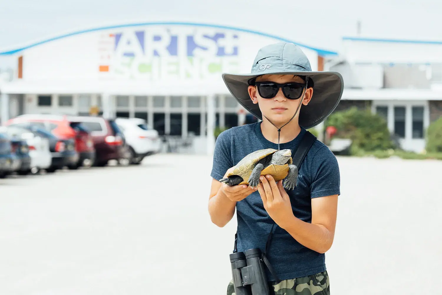 Child wearing a sun hat and sunglasses holds a turtle in front of the Long Beach Island Foundation of the Arts and Sciences building.