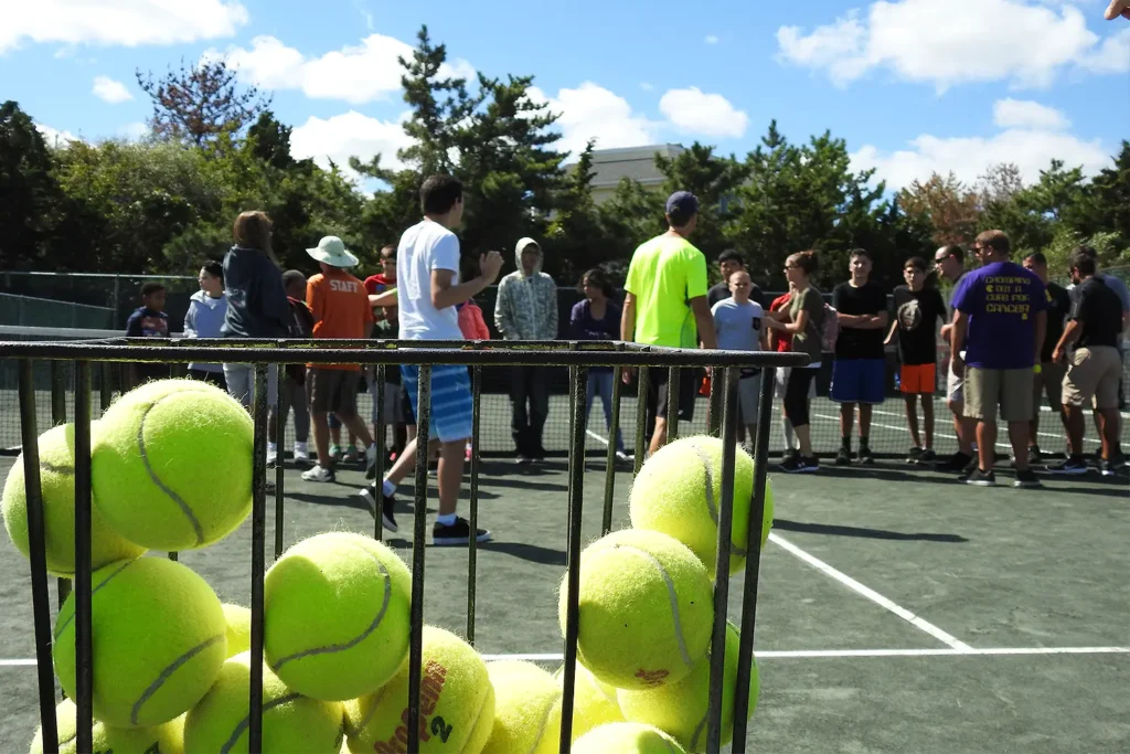 A basket of tennis balls in the foreground with a group of players gathered on the LBIF clay courts in the background.
