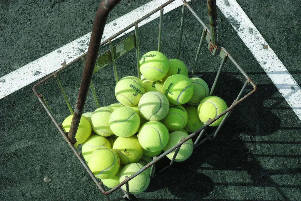 A metal basket filled with bright yellow tennis balls sits on the green clay court at LBIF.