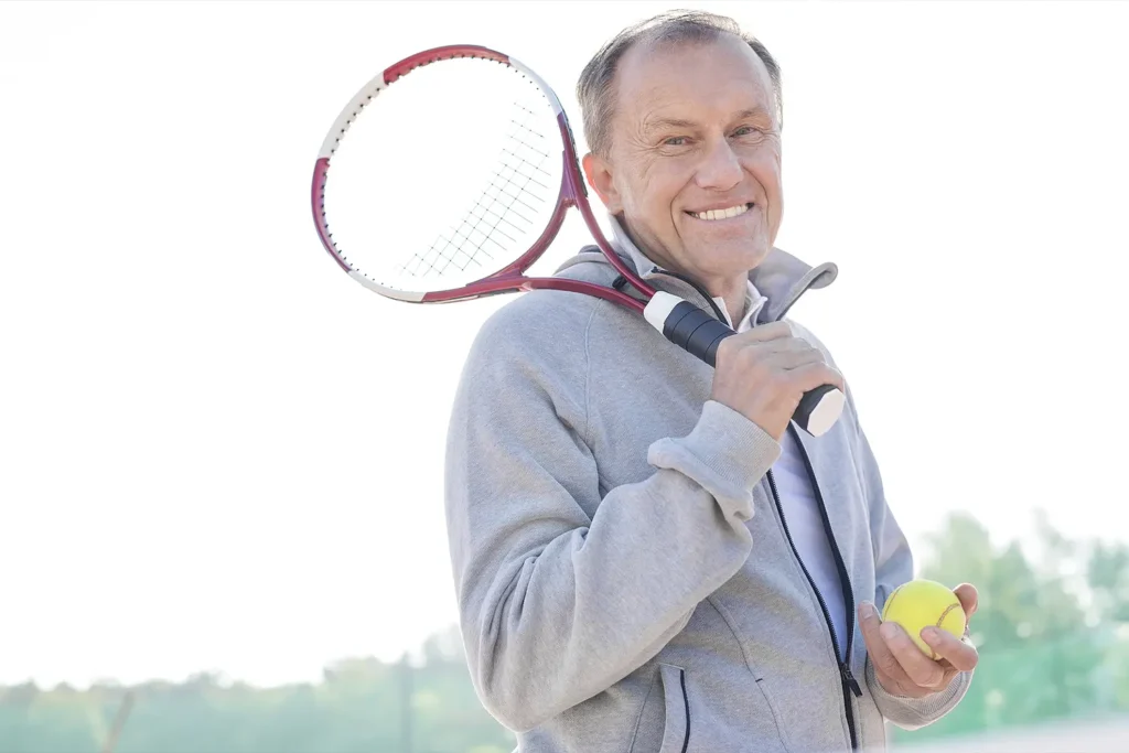 A smiling man holds a tennis racket over his shoulder and a tennis ball in his hand, standing outdoors on a bright day.