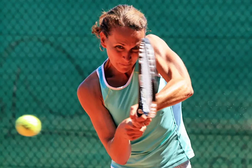 A focused tennis player follows through on a powerful backhand shot, the ball visible mid-air against a green fence backdrop.