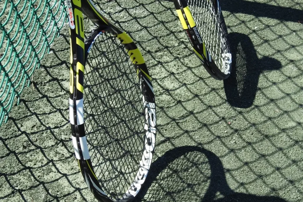 Two tennis rackets lean against a green chain-link fence, their shadows stretching across the clay court in the sunlight.