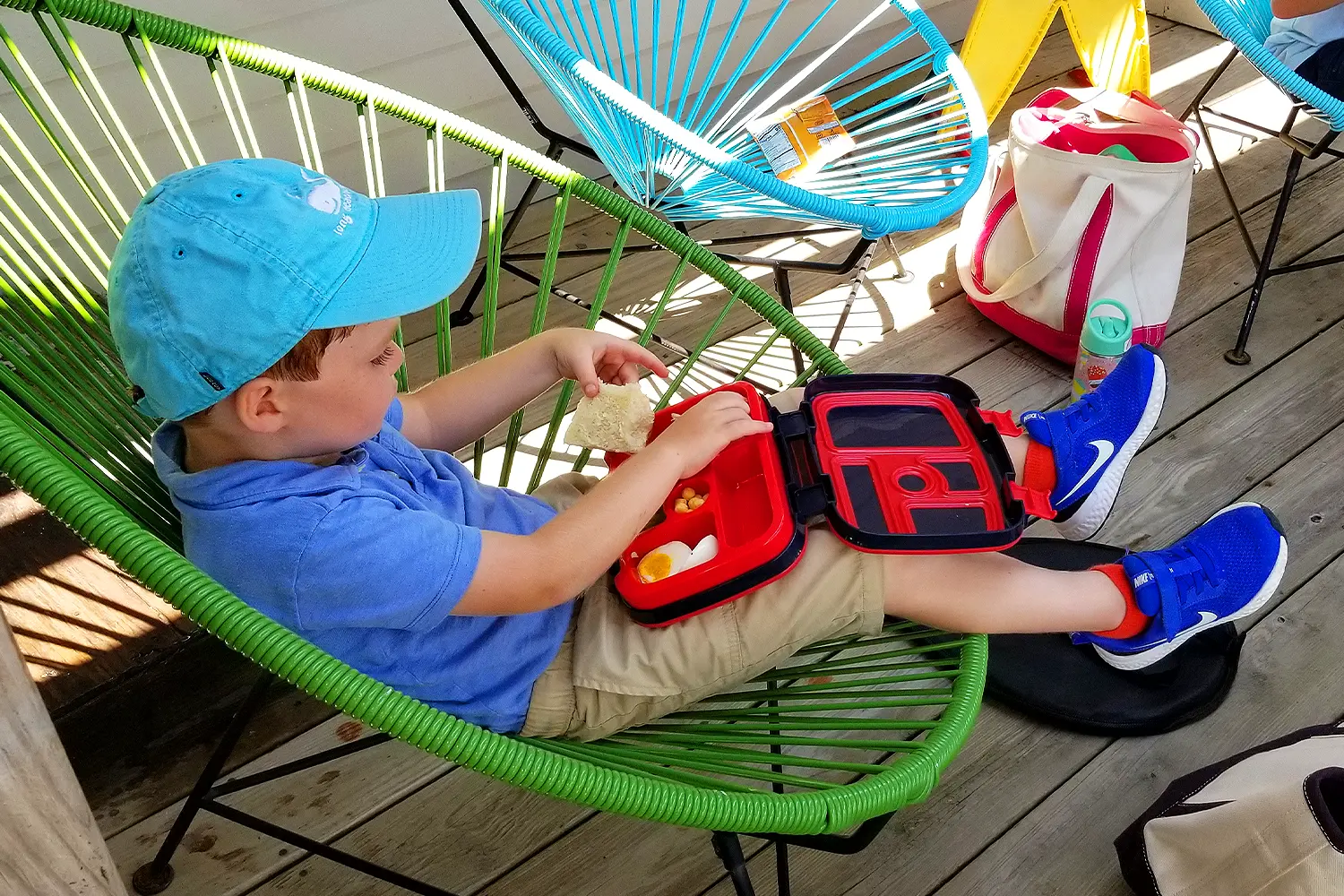 A young camper relaxes in a green chair while eating lunch from a red bento box at LBIF Summer Camp.