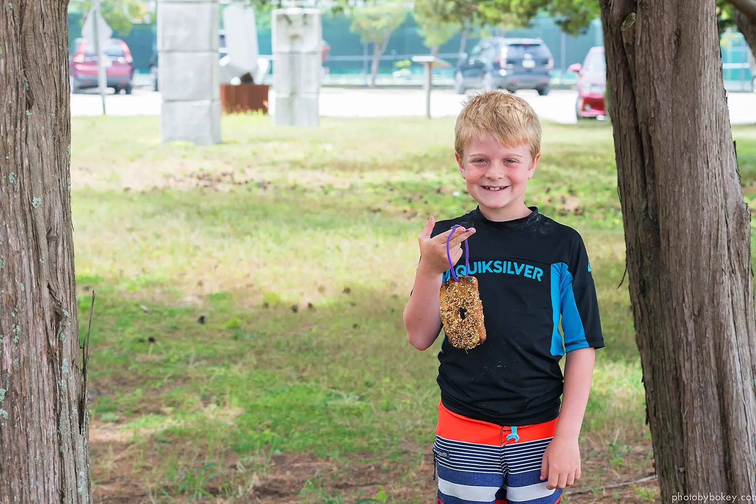 A smiling camper stands outdoors holding a handmade bird feeder covered in seeds at LBIF’s Summer Camp.