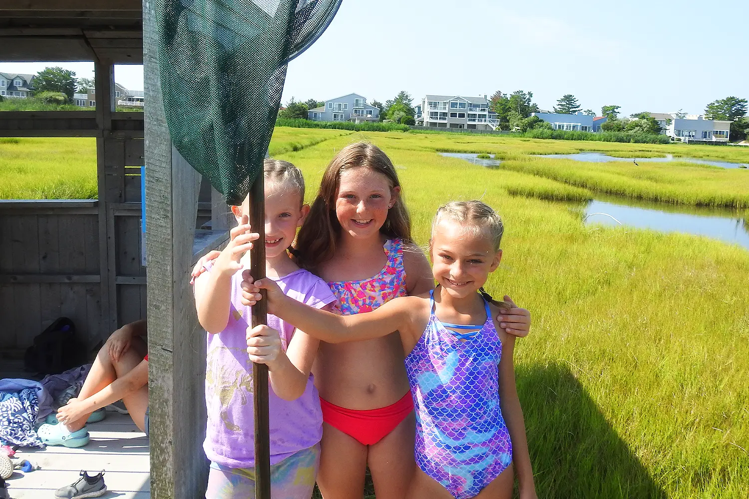 Three campers in swimsuits smile while holding a large fishing net in front of the salt marsh at LBIF Summer Camp.