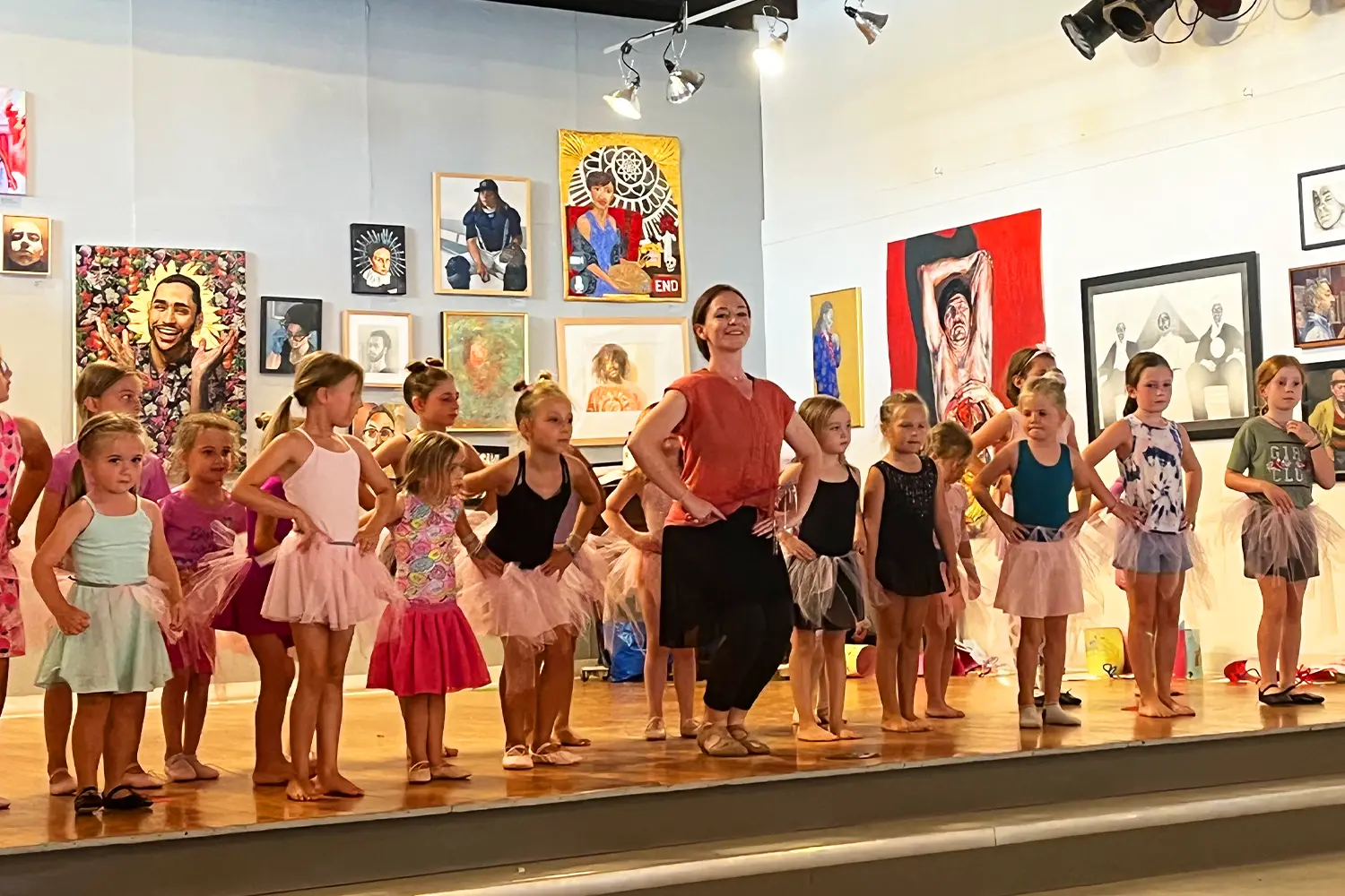 A dance instructor leads a group of young campers in ballet practice on stage at LBIF, surrounded by colorful artwork on the gallery walls.