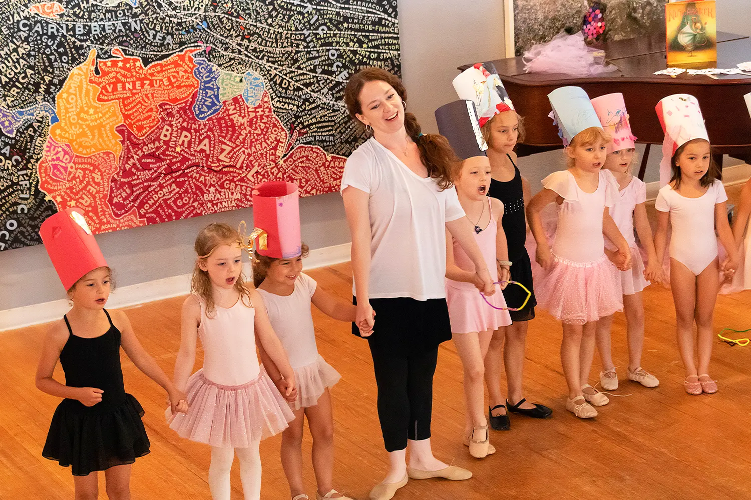 A group of young ballerinas in pink and black tutus hold hands with their instructor during a dance performance, wearing handmade paper hats and smiling in front of a colorful mural.