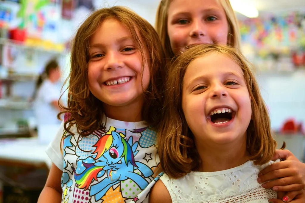 Three young girls smiling and laughing together in an art classroom filled with bright colors and creative energy.