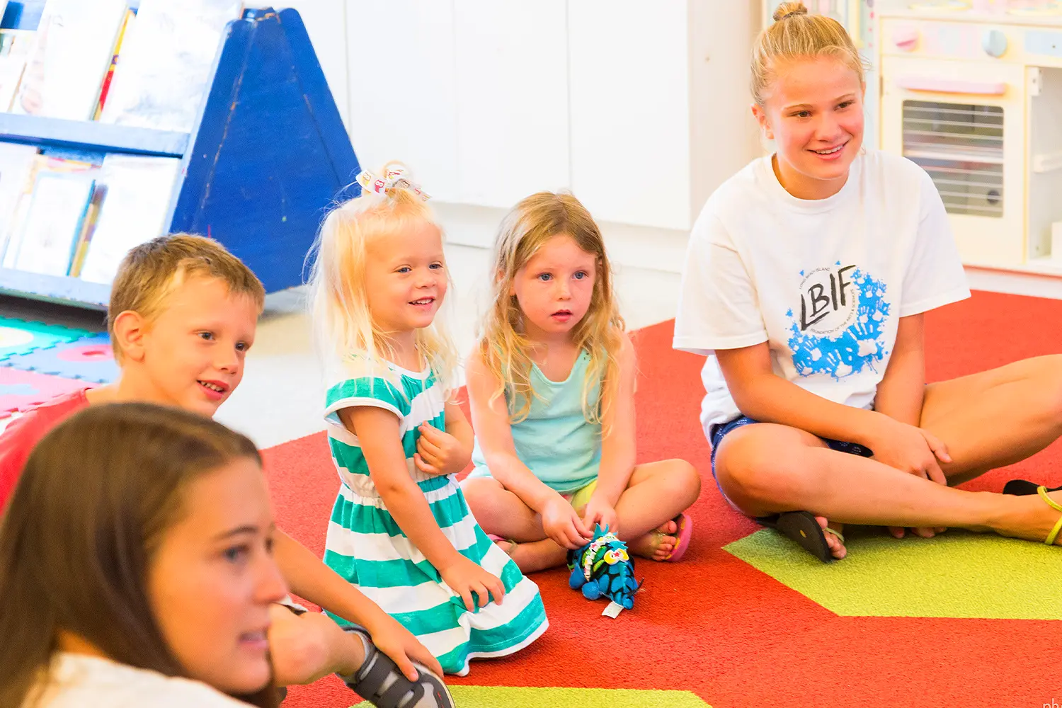 A group of young children sit on a colorful classroom rug, smiling and listening attentively to an instructor wearing an LBIF t-shirt.