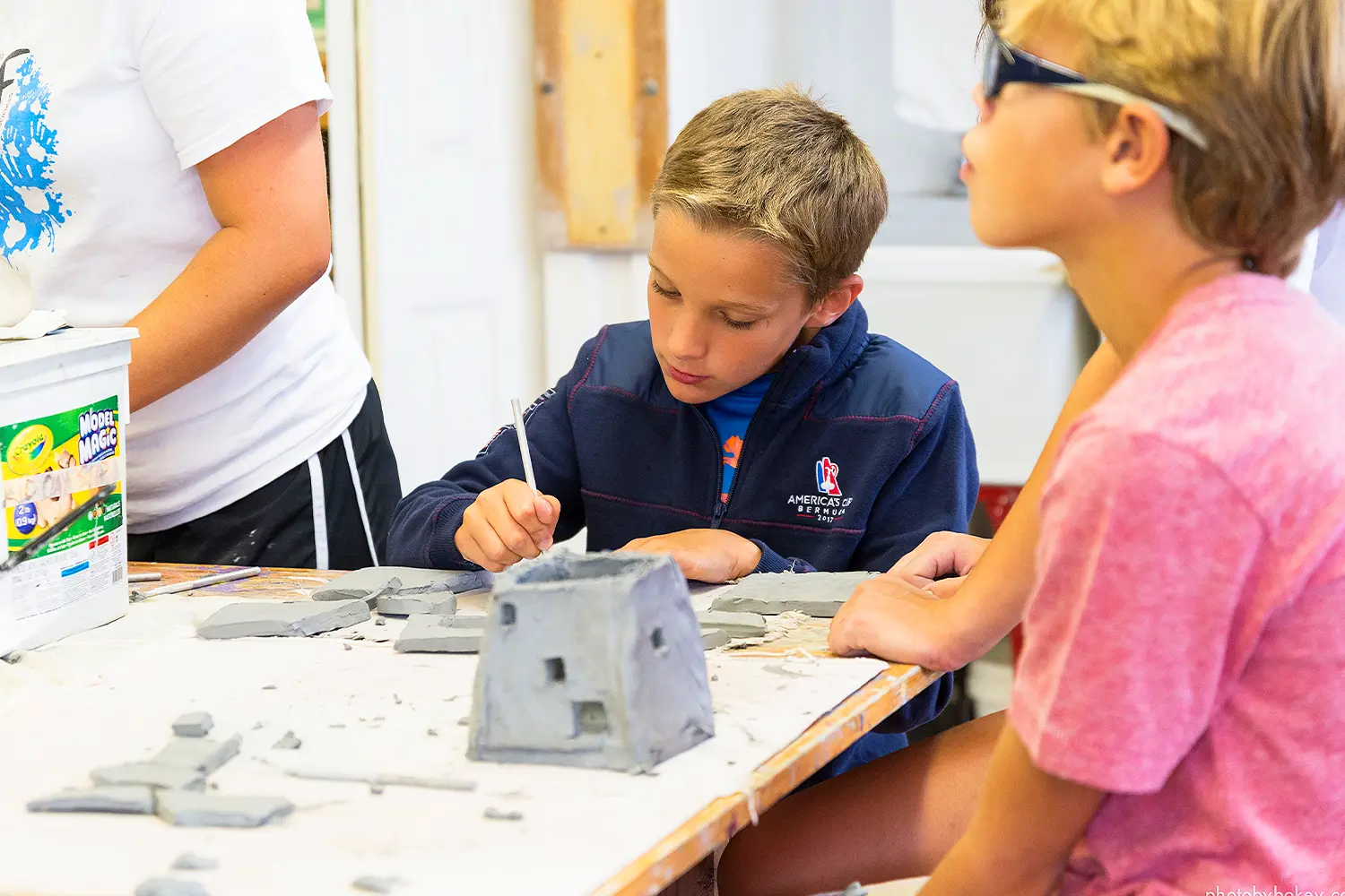A young camper focuses intently while sculpting with clay during an art class, surrounded by tools and pieces of gray clay on the table.