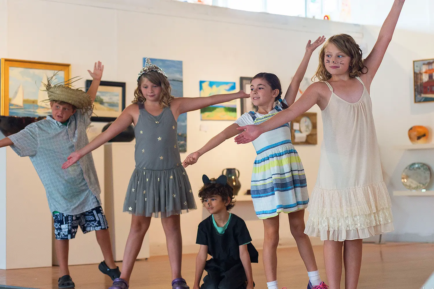 A group of campers in costume perform a play inside an art gallery, posing with arms outstretched in unison.