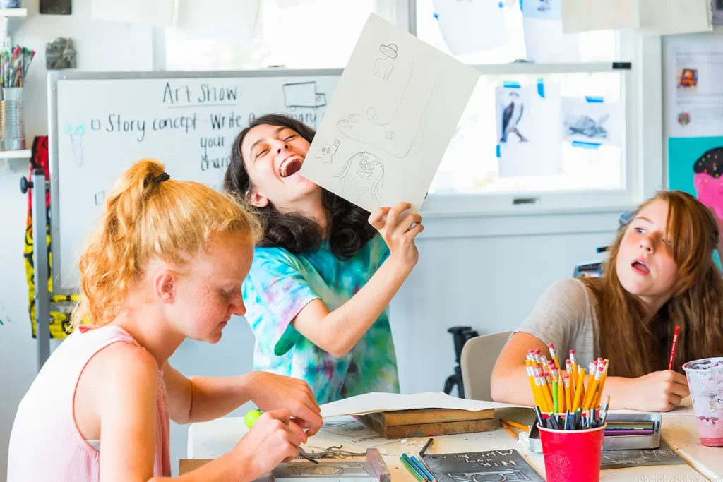 Three campers sit at a table inside an art studio, with one camper holding up a drawing and laughing while others work on their projects.