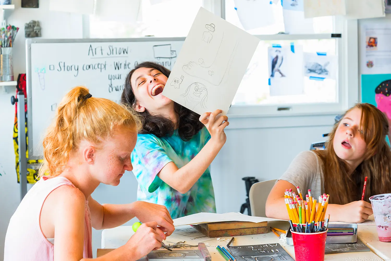 Three campers sit at a table inside an art studio, with one camper holding up a drawing and laughing while others work on their projects.