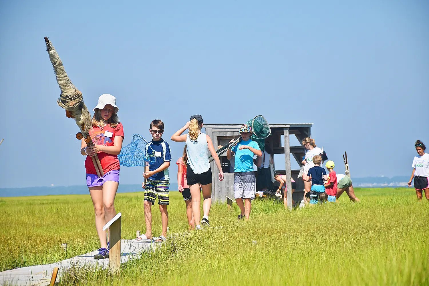 Children walk along a marsh boardwalk carrying nets and field tools during an outdoor environmental program.