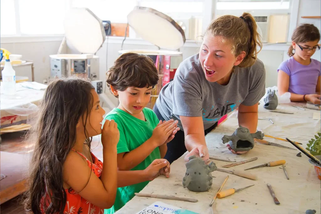 A camp instructor guides children as they shape clay sculptures at a table inside a ceramics studio.