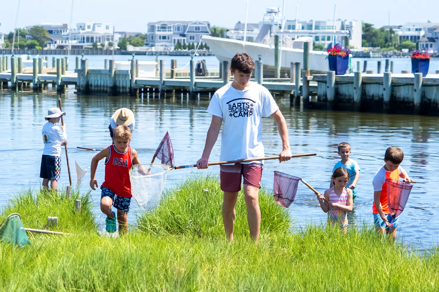 Camp counselor leads children through marsh grass while campers use nets to explore shallow bay water.