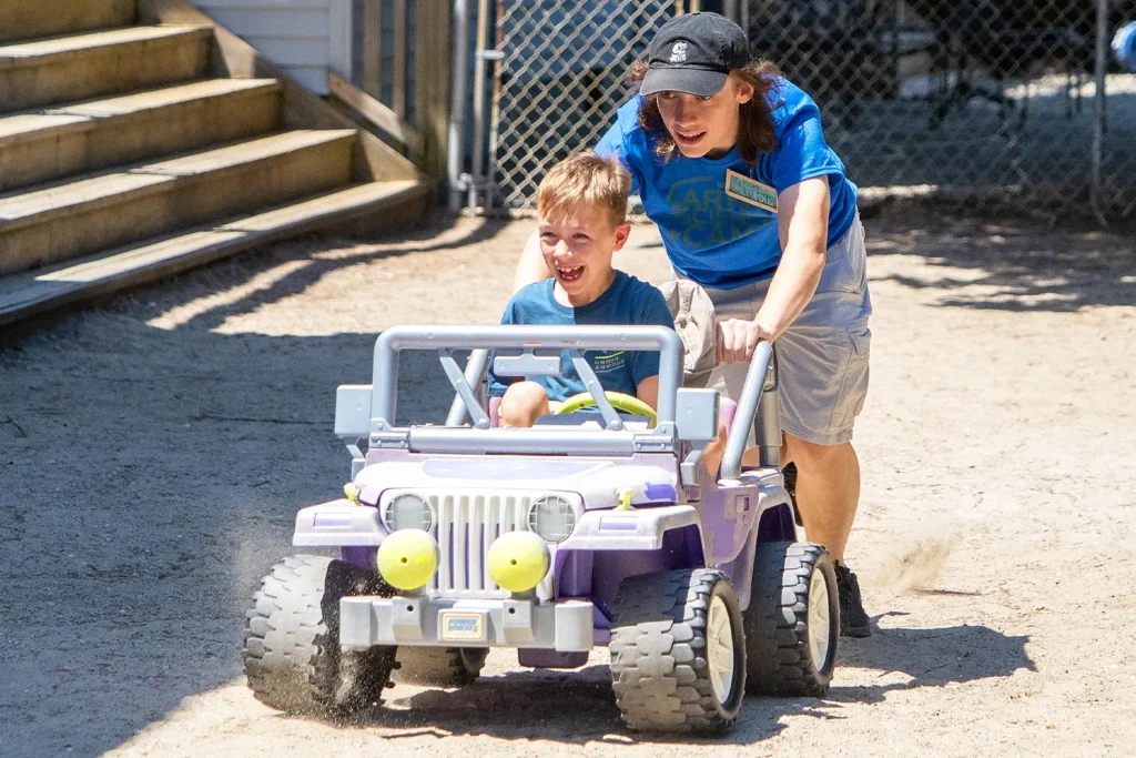 Camp counselor pushing a smiling child in a small toy jeep on a sandy outdoor play area.