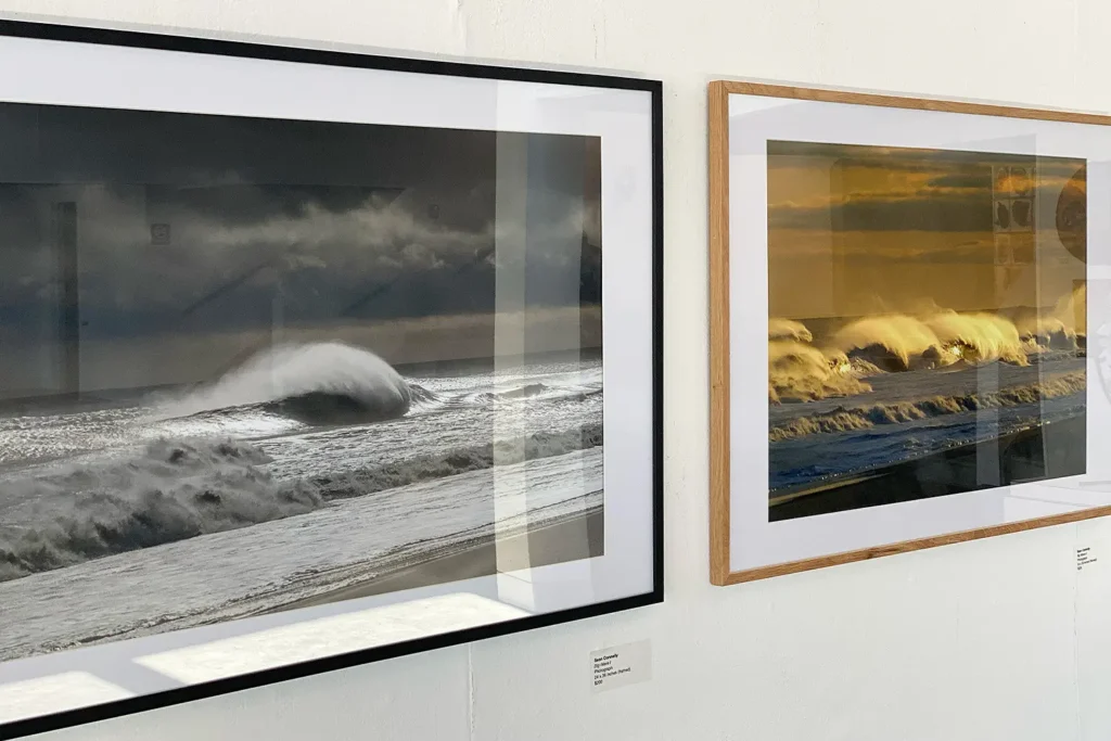 Two framed ocean wave photographs displayed side by side on a white gallery wall, one in grayscale and the other bathed in golden light.