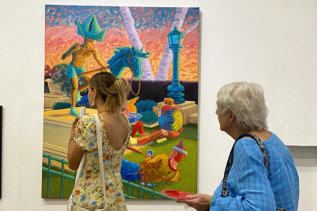 Two women view a colorful, surreal painting featuring headless figures, a horse, and a lamppost under a vivid orange and blue sky.