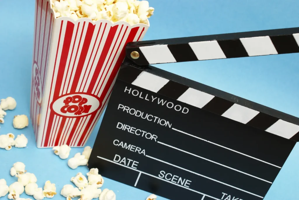 Popcorn in a red-and-white striped container beside a black film clapperboard on a light blue background, symbolizing movie screenings.