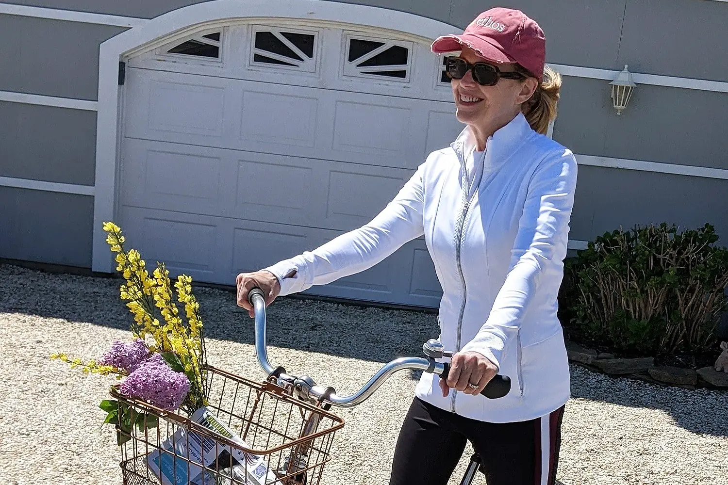 A smiling woman in a white jacket and red cap rides a bicycle with flowers and a rolled yoga mat in the basket.