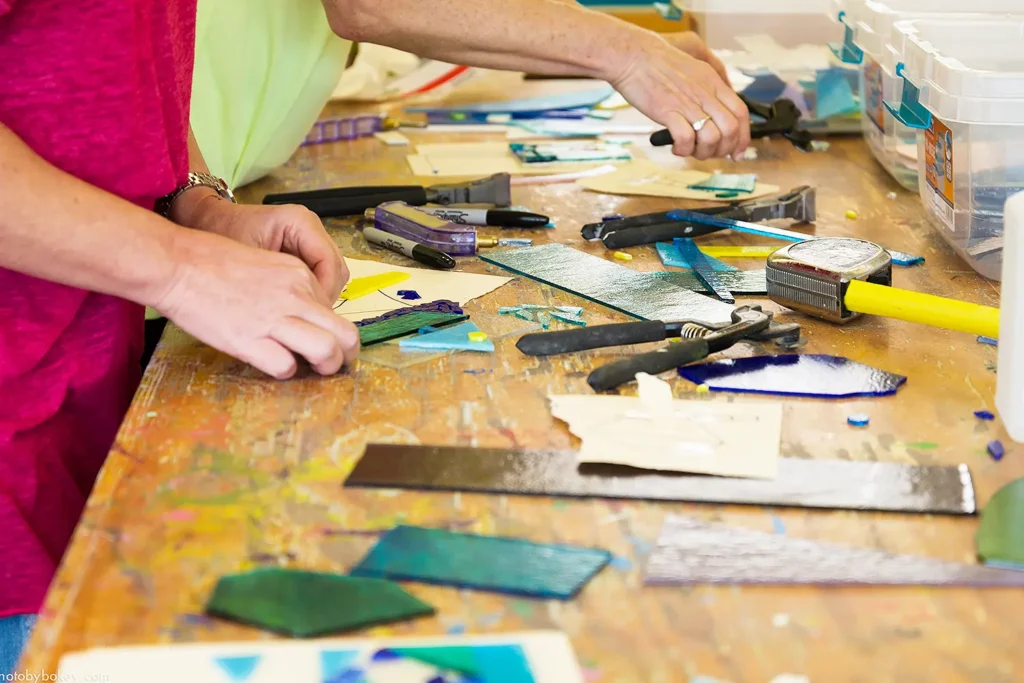 Close-up of hands arranging colorful glass pieces and tools on a worktable during an adult glass mosaic workshop at the Long Beach Island Foundation of the Arts & Sciences.
