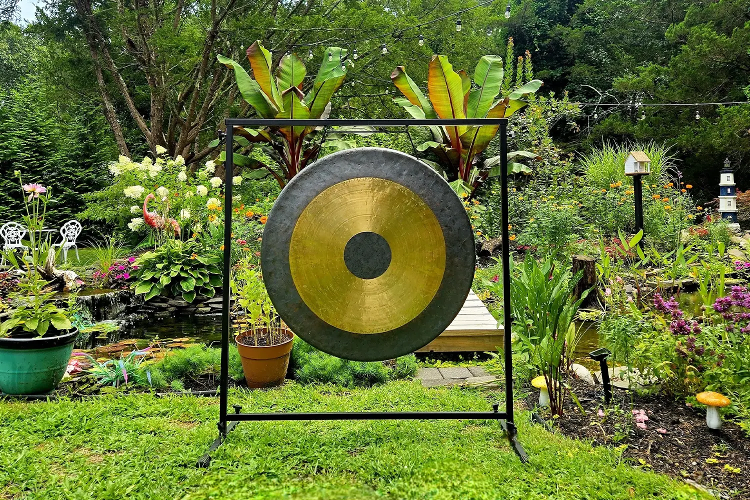 A large golden gong stands in the center of a lush garden surrounded by colorful flowers, plants, and a small pond.