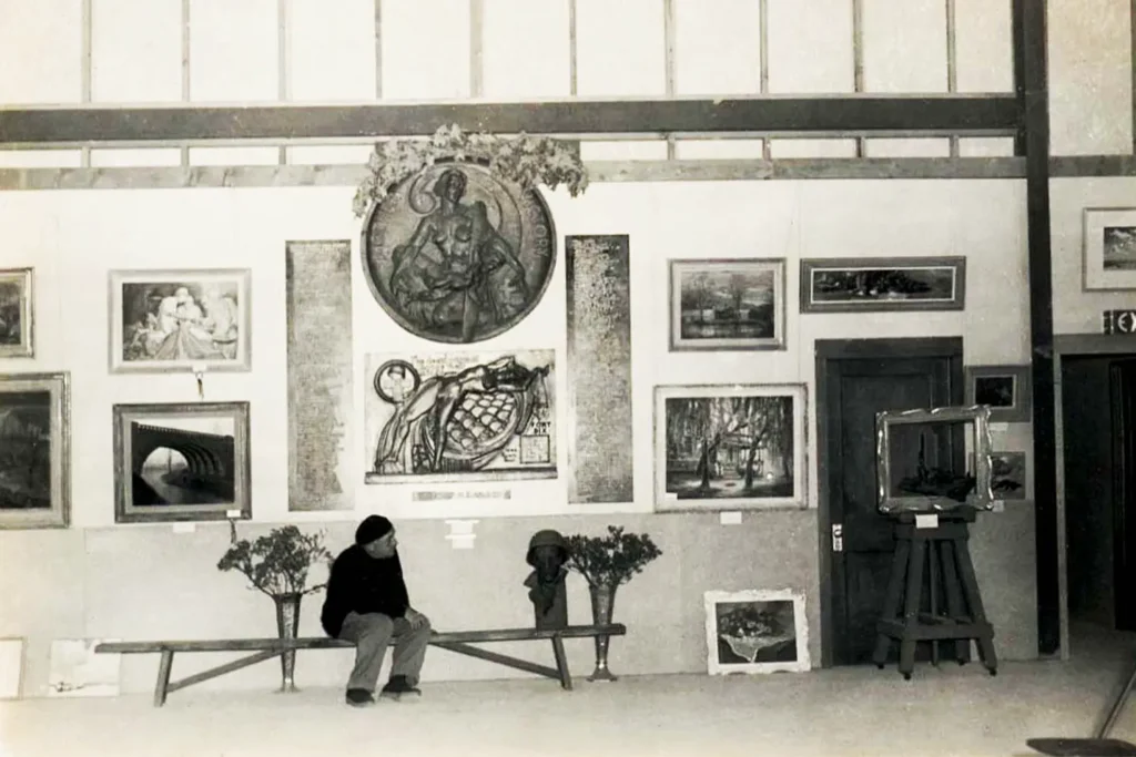 Black-and-white photograph of an early LBIF gallery interior, with framed artworks covering the walls and a seated artist resting on a bench beneath the exhibition display.