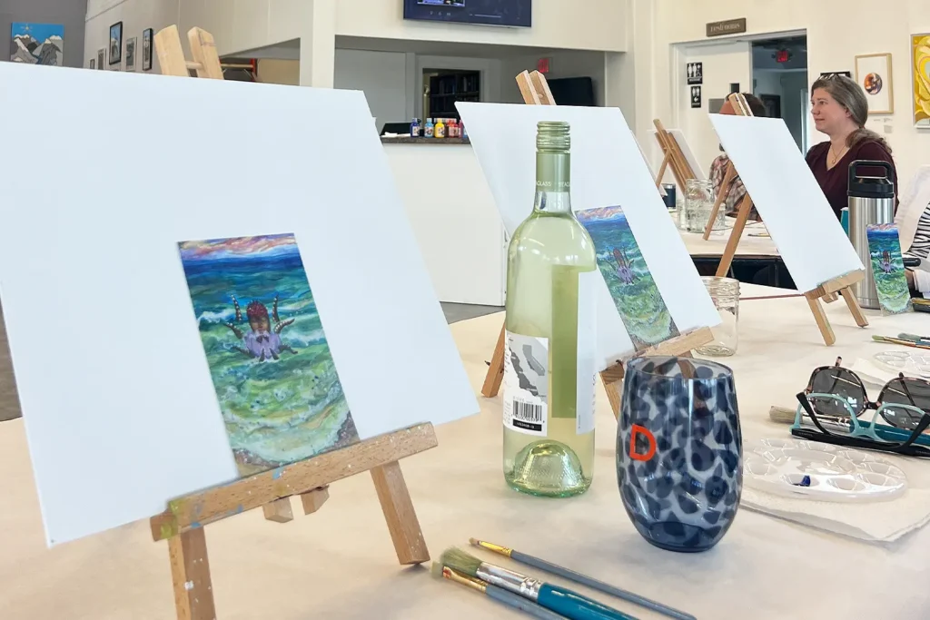 Small canvases on tabletop easels are arranged for a paint-and-sip workshop, with paintbrushes, a wine bottle, and glasses set on the table as participants work in the background at LBIF.