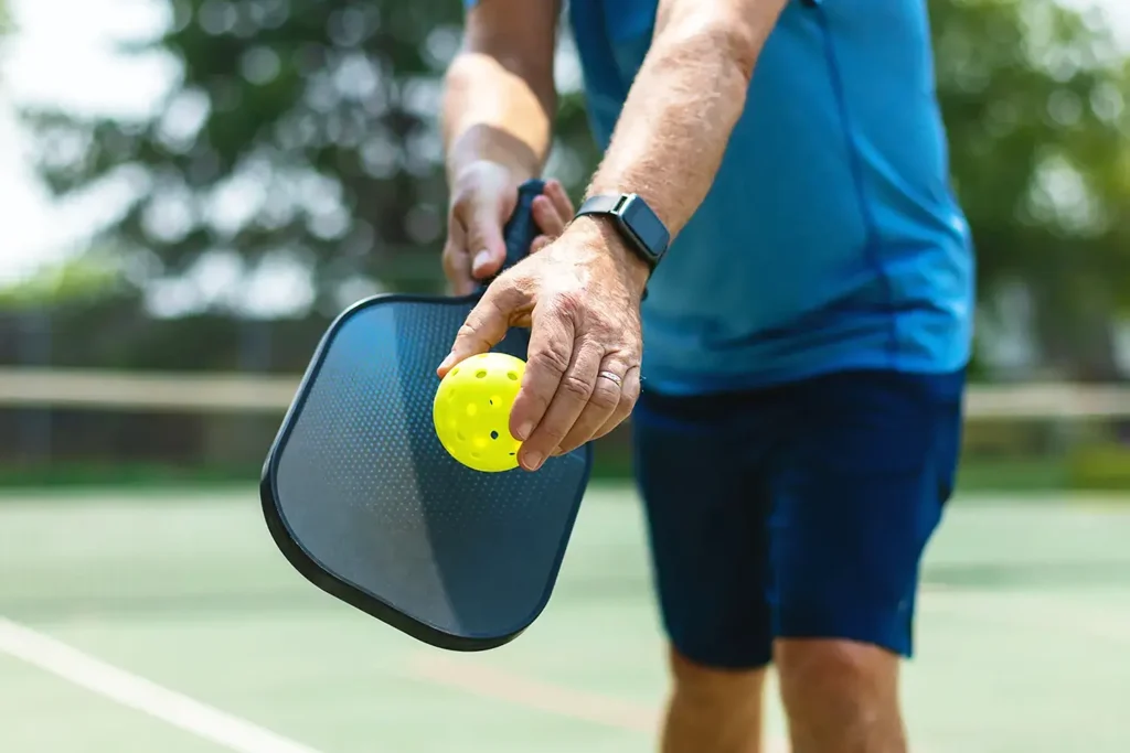 Close-up of a person about to serve in a pickleball game, holding a yellow pickleball above a black paddle on an outdoor court.