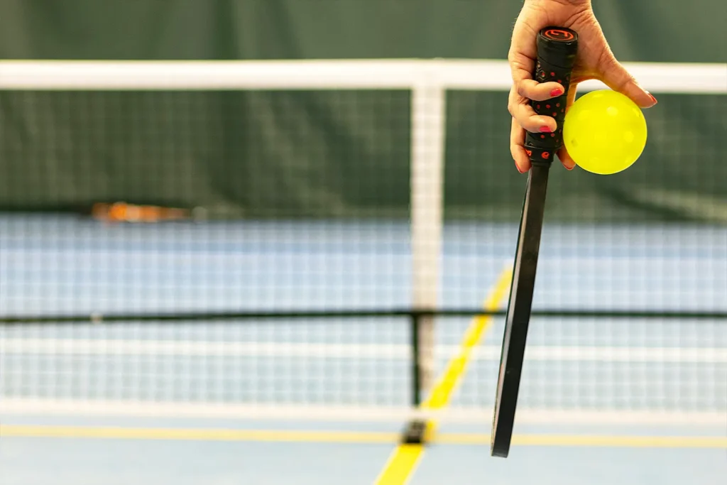 A person’s hand holding a yellow pickleball and paddle, preparing to serve on an indoor court with a net in the background.