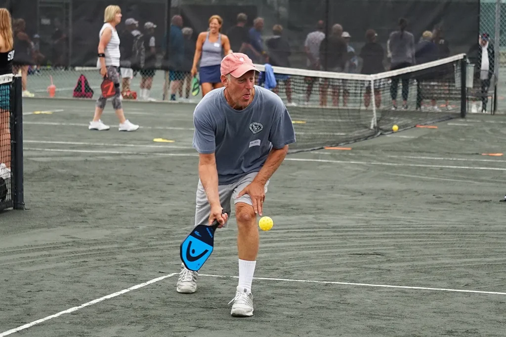A man wearing a gray shirt and pink cap focuses as he returns a pickleball shot on an outdoor court at LBIF, with other players and nets visible in the background.