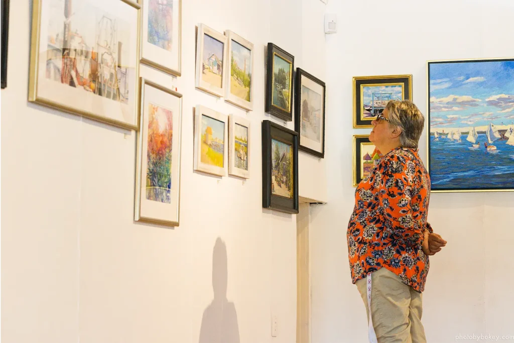 An adult visitor stands in a gallery viewing a wall of framed landscape and coastal paintings during the Plein Air Plus exhibition at LBIF.