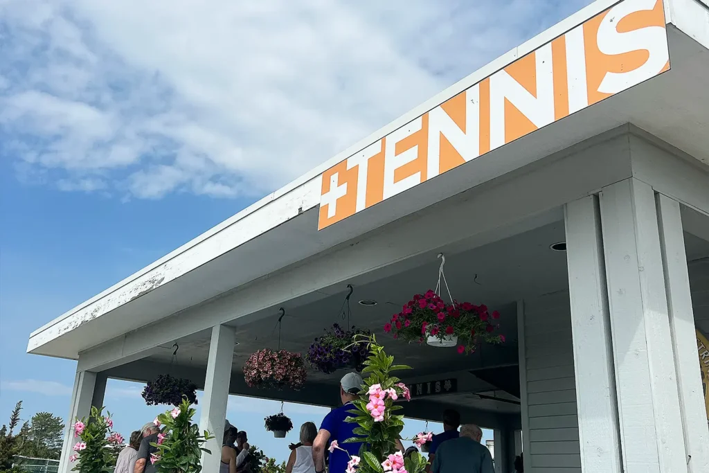 Upward view of LBIF’s Tennis Pavilion showing the bold orange “+Tennis” sign along the roofline, hanging flower baskets, and visitors gathered beneath the covered structure on a partly cloudy day.