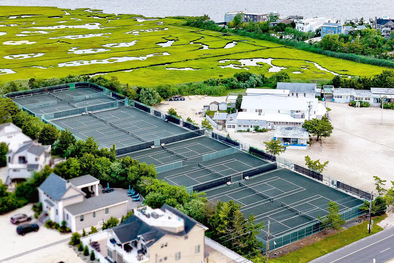 Aerial view of LBIF’s tennis courts, showing multiple outdoor courts bordered by trees, nearby buildings, and expansive coastal marshland stretching toward the bay.