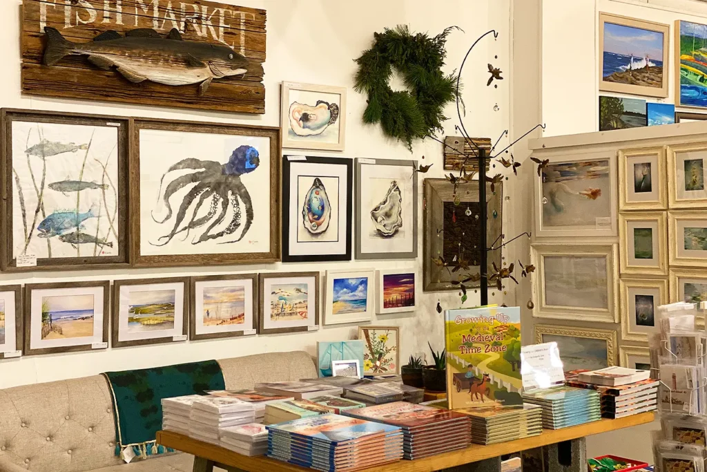 Art prints, framed coastal artwork, and stacks of books displayed on a table and gallery wall at a holiday market.