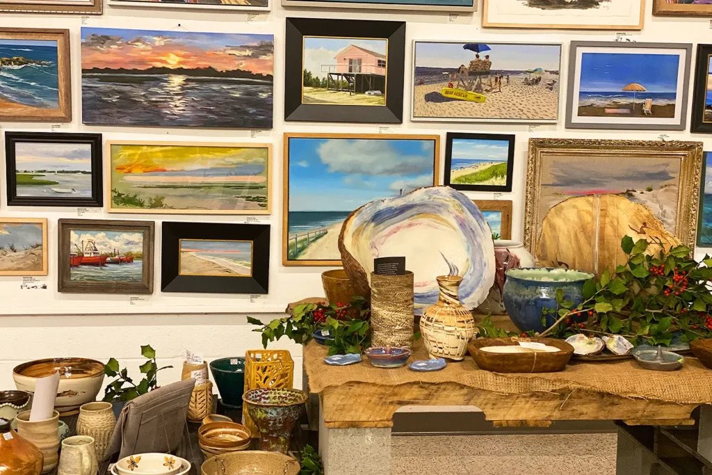 Gallery wall filled with framed coastal paintings above a table displaying handmade ceramics and decorative bowls.