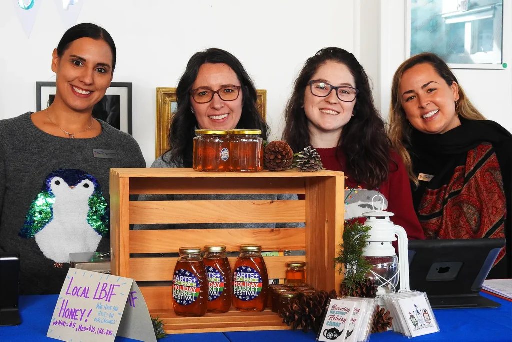 Four smiling women stand behind a vendor table displaying jars of local honey and festive décor at a Holiday Market Festival.