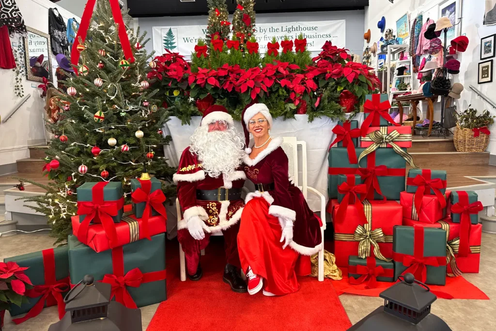 Santa Claus and Mrs. Claus sit together on a red carpet surrounded by wrapped gifts, poinsettias, and decorated Christmas trees at a holiday market.