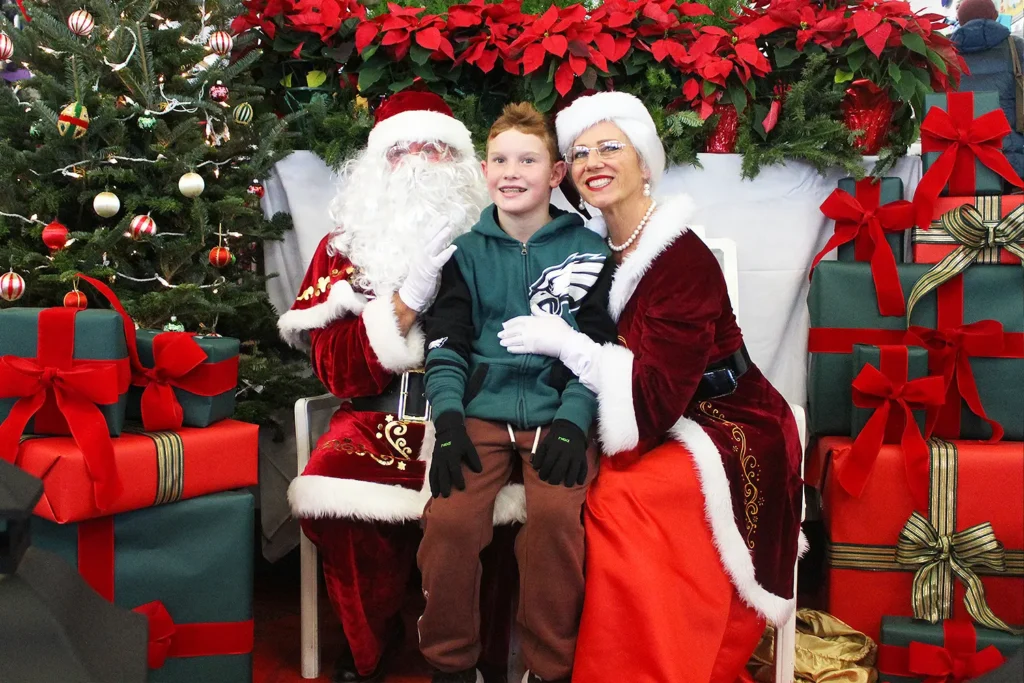 A smiling child sits between Santa and Mrs. Claus on a chair, surrounded by poinsettias, a decorated Christmas tree, and stacks of wrapped gifts in a festive holiday photo area.